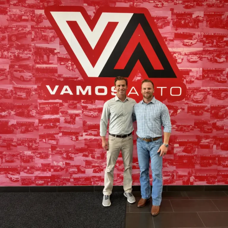 Two men posing in front of a red wall with the Vamos Auto logo and car collage background.
